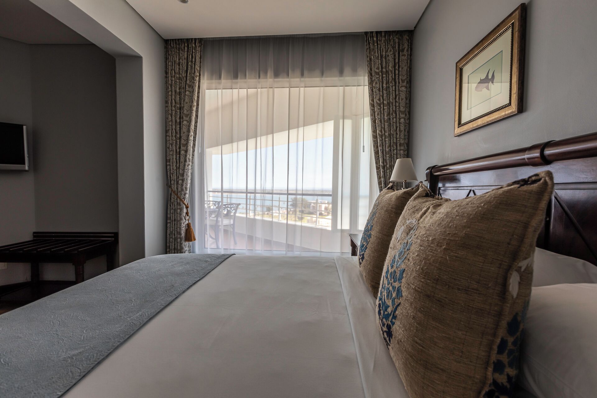 A hotel room with a large bed, a view of the city through the window, a small table, and a framed picture on the wall.
