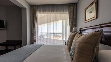 A hotel room with a large bed, a view of the city through the window, a small table, and a framed picture on the wall.