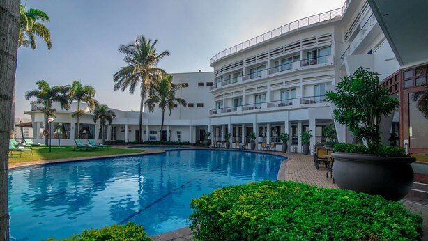 A modern hotel with a swimming pool, palm trees, and a multi-story building with balconies.