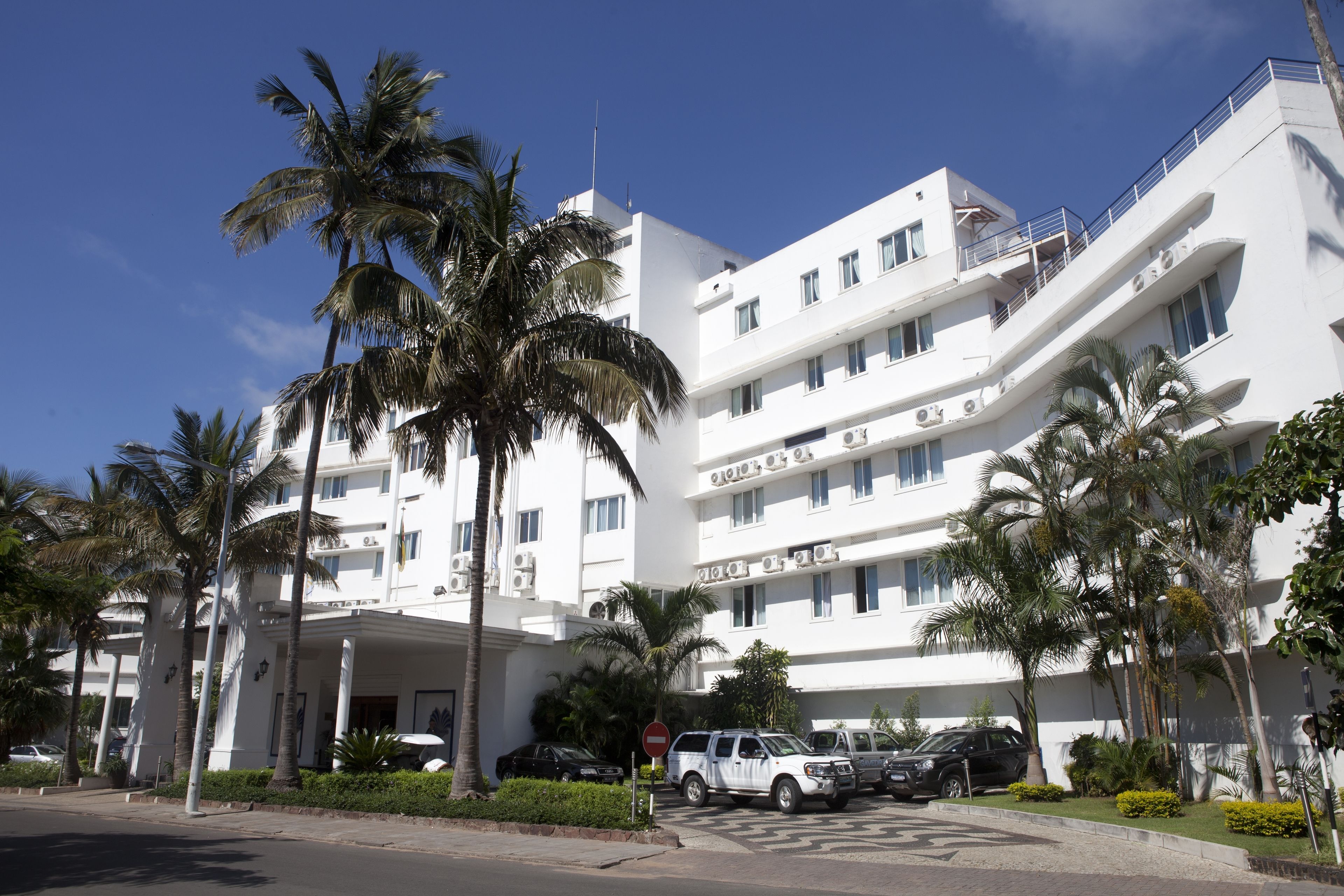 A multi-story hotel with a white facade, palm trees, and a clear blue sky.