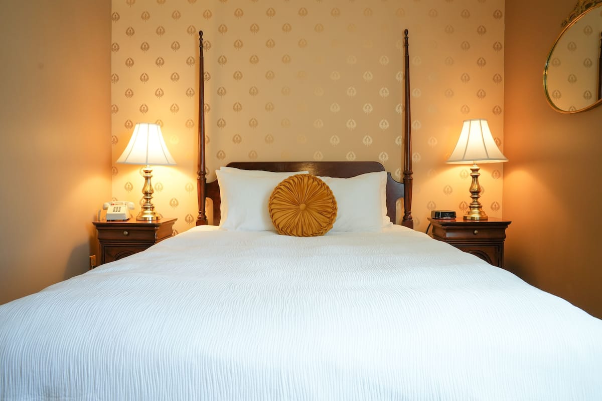 Una cama bien hecha con un cojín decorativo grande y redondo, dos mesitas de noche con lámparas y un espejo en la pared.