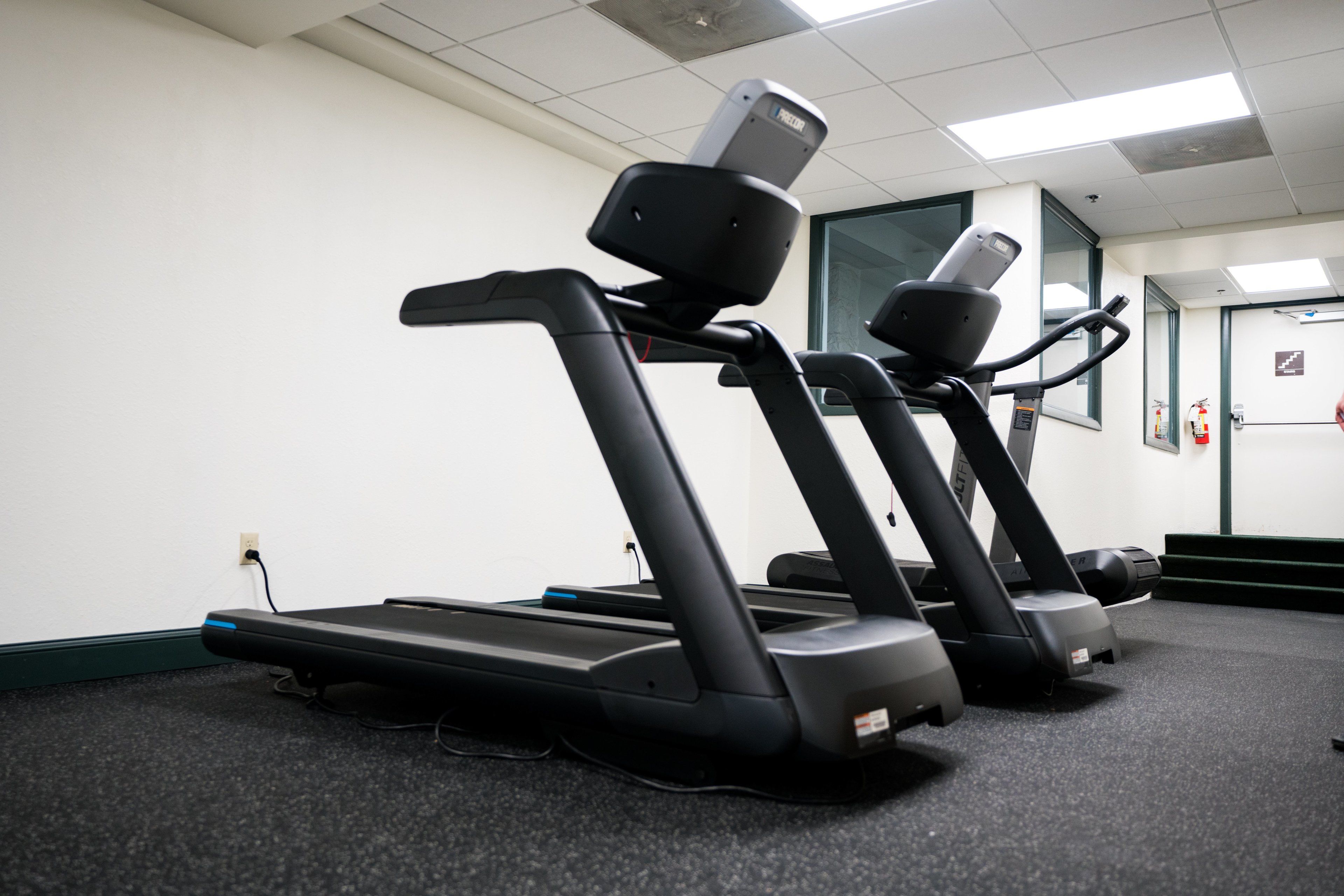 A gym with three treadmills aligned against a wall, a fire extinguisher mounted on the wall, and a door with a sign.