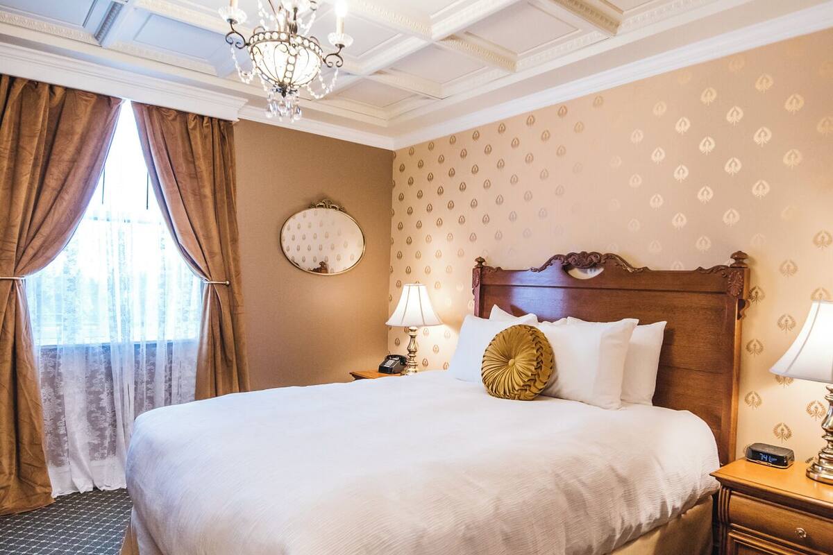 Una habitación de hotel con una cama grande, una araña, un espejo redondo y una mesita de noche con lámpara.