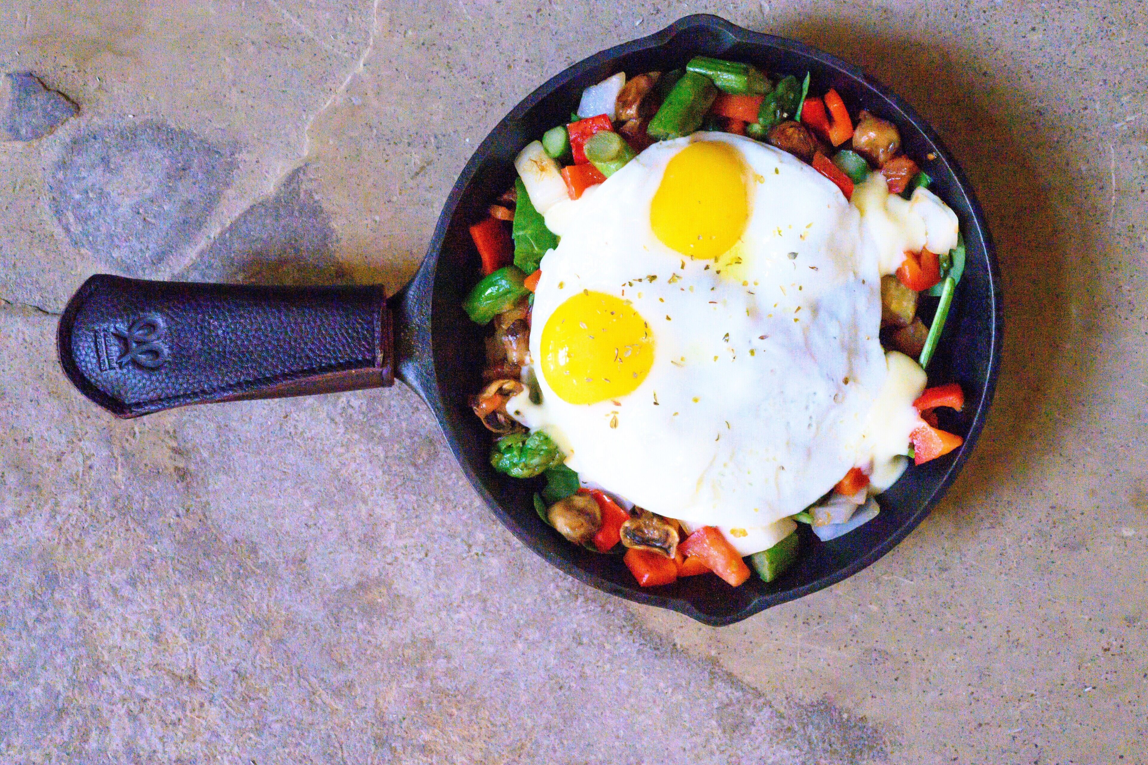A skillet with eggs and vegetables on a stone surface.