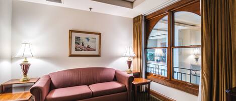 A hotel room with a red sofa, two side tables with lamps, a framed picture on the wall, and a large window with curtains.