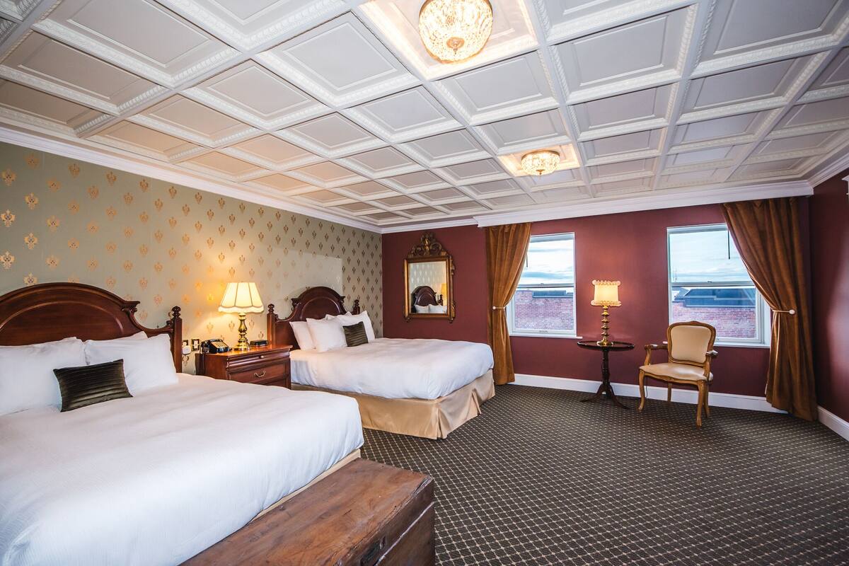 Habitación de hotel con dos camas, cabecera de madera, mesita de noche con lámpara, una silla y vista al mar a través de una ventana.