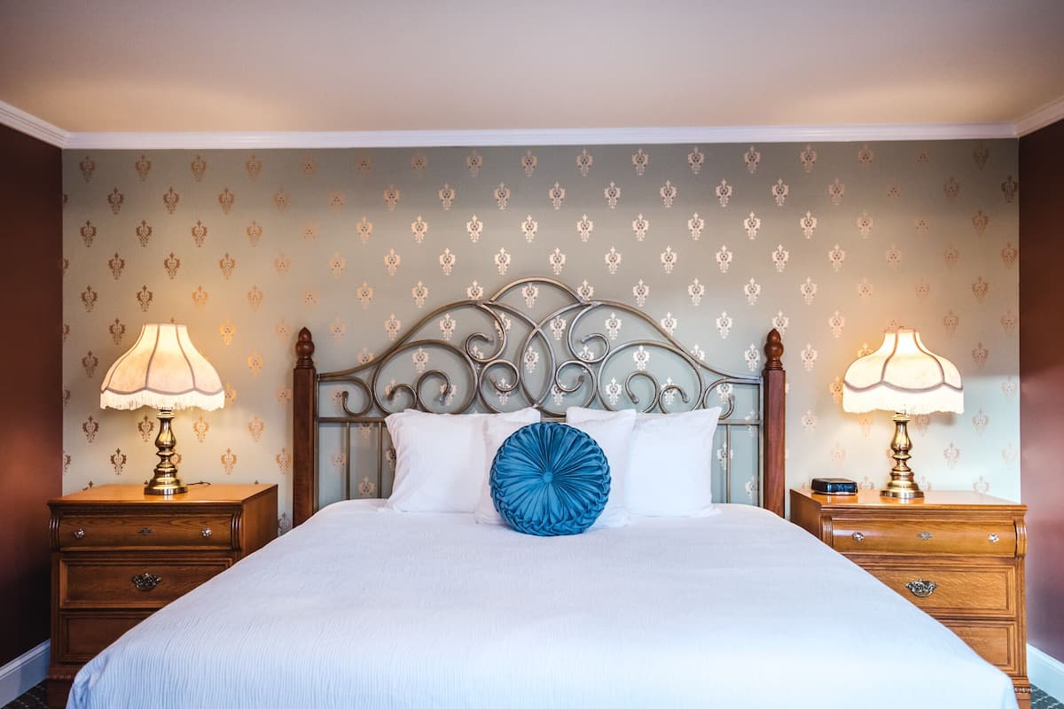 Una cama bien hecha con un cabecero decorativo, dos mesitas de noche con lámparas y un papel tapiz estampado.