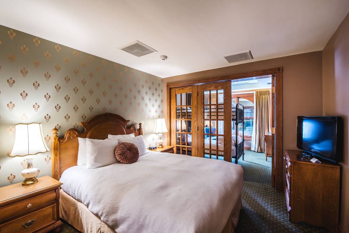 Habitación de hotel con una cama grande, cabecera de madera, un televisor y vistas al exterior por una puerta de cristal.