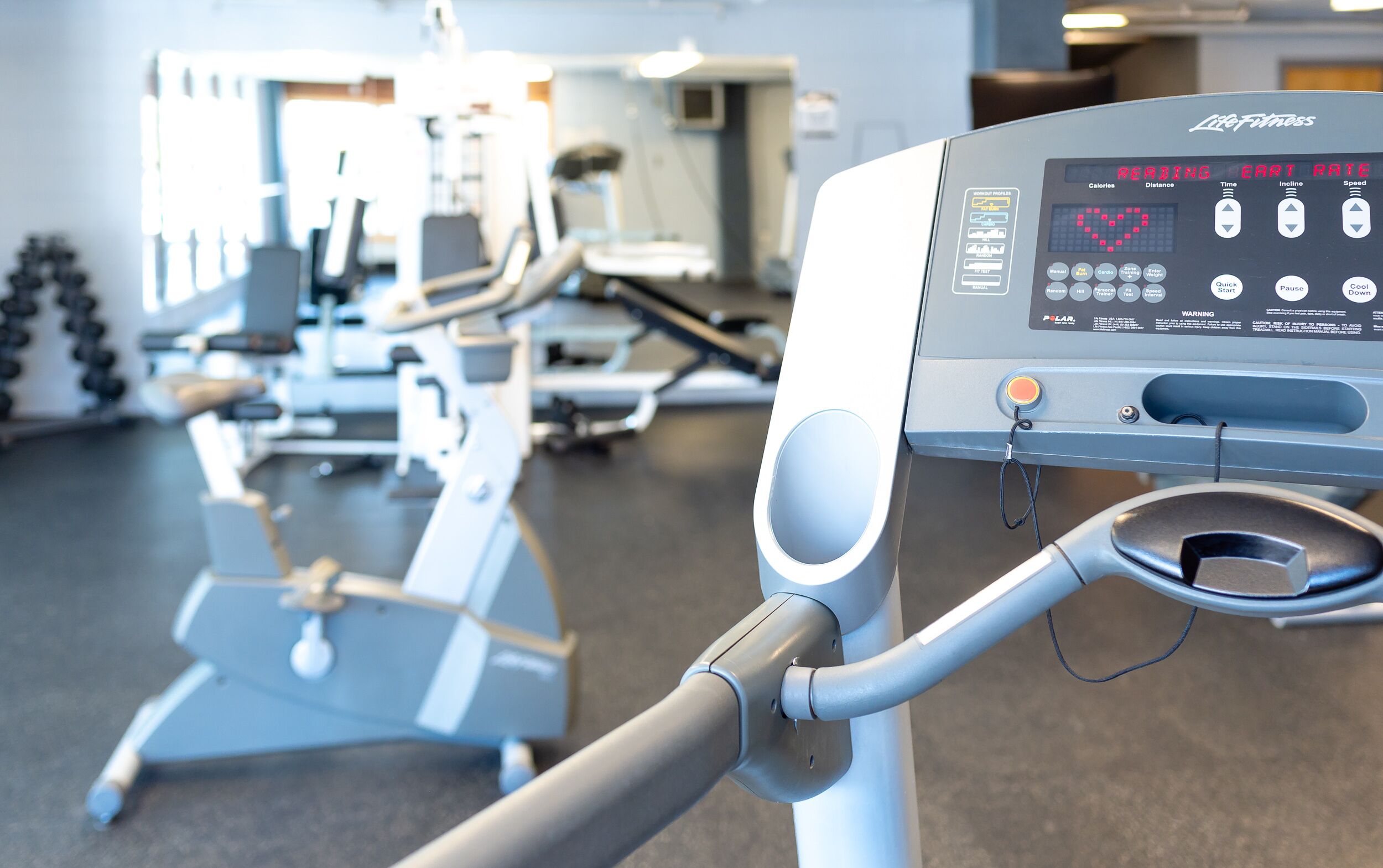 Ein Laufband mit digitalem Display, das Herzfrequenz und andere Trainingsdaten anzeigt, in einem Fitnessstudio mit verschiedenen Geräten.