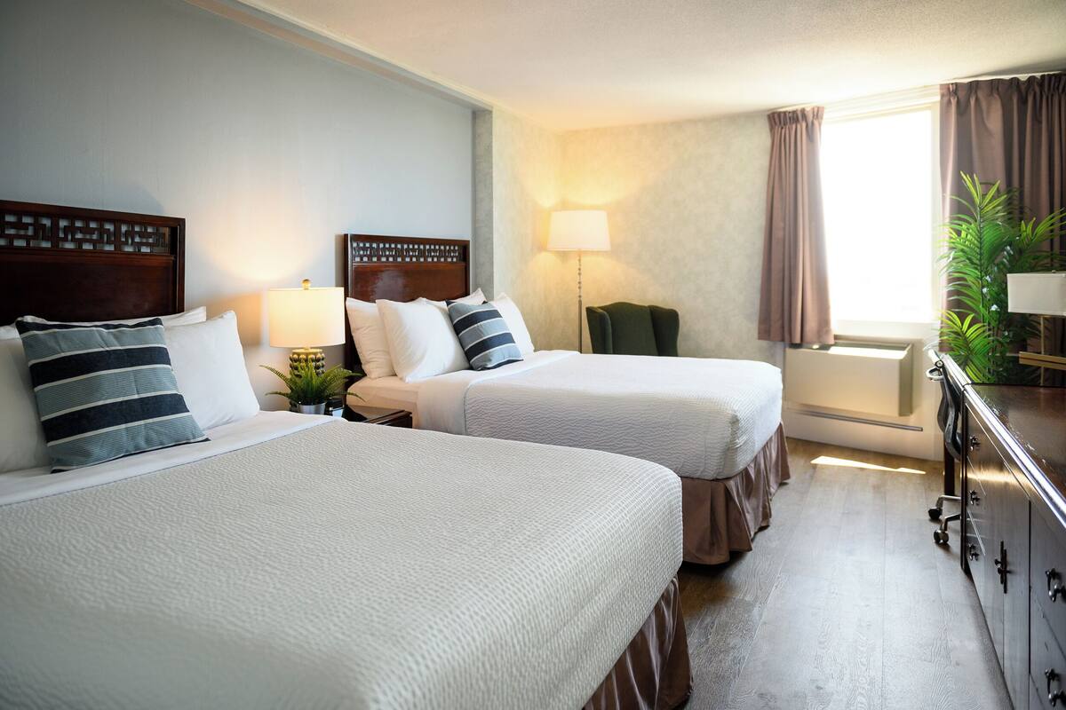 A hotel room with two beds, a desk, a lamp, a plant, and a window with curtains.