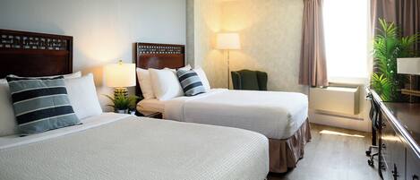 Ein Hotelzimmer mit zwei Betten, einem Schreibtisch, einer Lampe, einer Pflanze und einem Fenster mit Vorhängen.