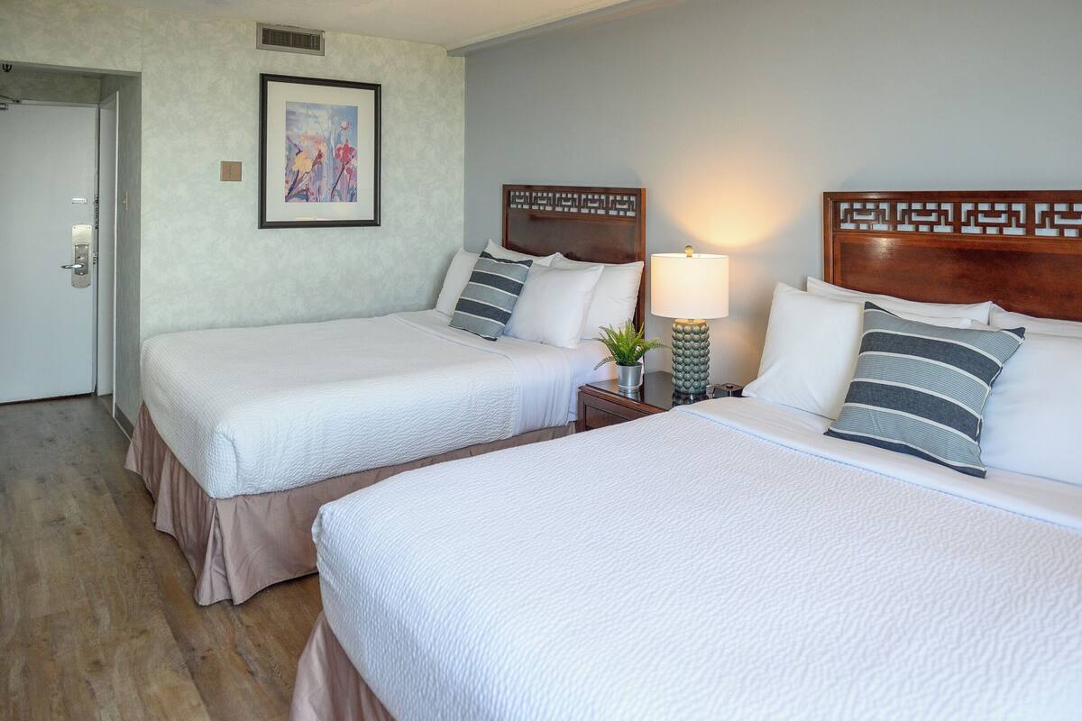 A hotel room with two beds, a nightstand with a lamp, a framed picture on the wall, and a door to the left.