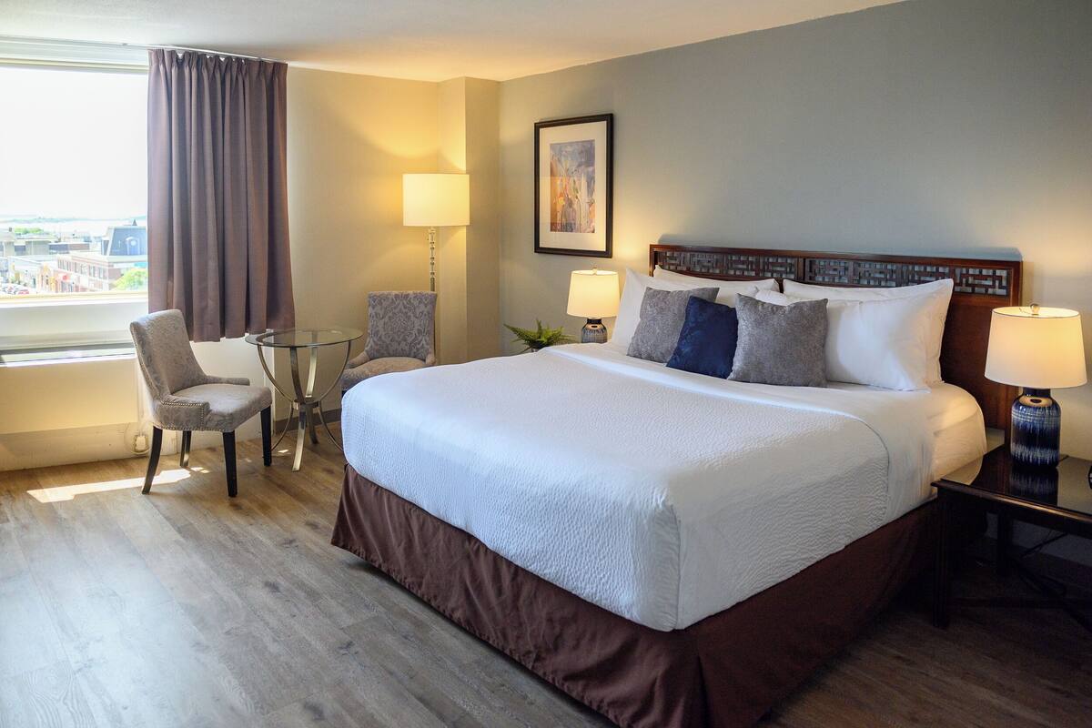 A hotel room with a large bed, two bedside tables with lamps, a chair, a small table, and a window with curtains.