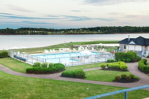 A swimming pool with lounge chairs and umbrellas, surrounded by well-maintained lawns and bushes, with a lake and trees in the background.