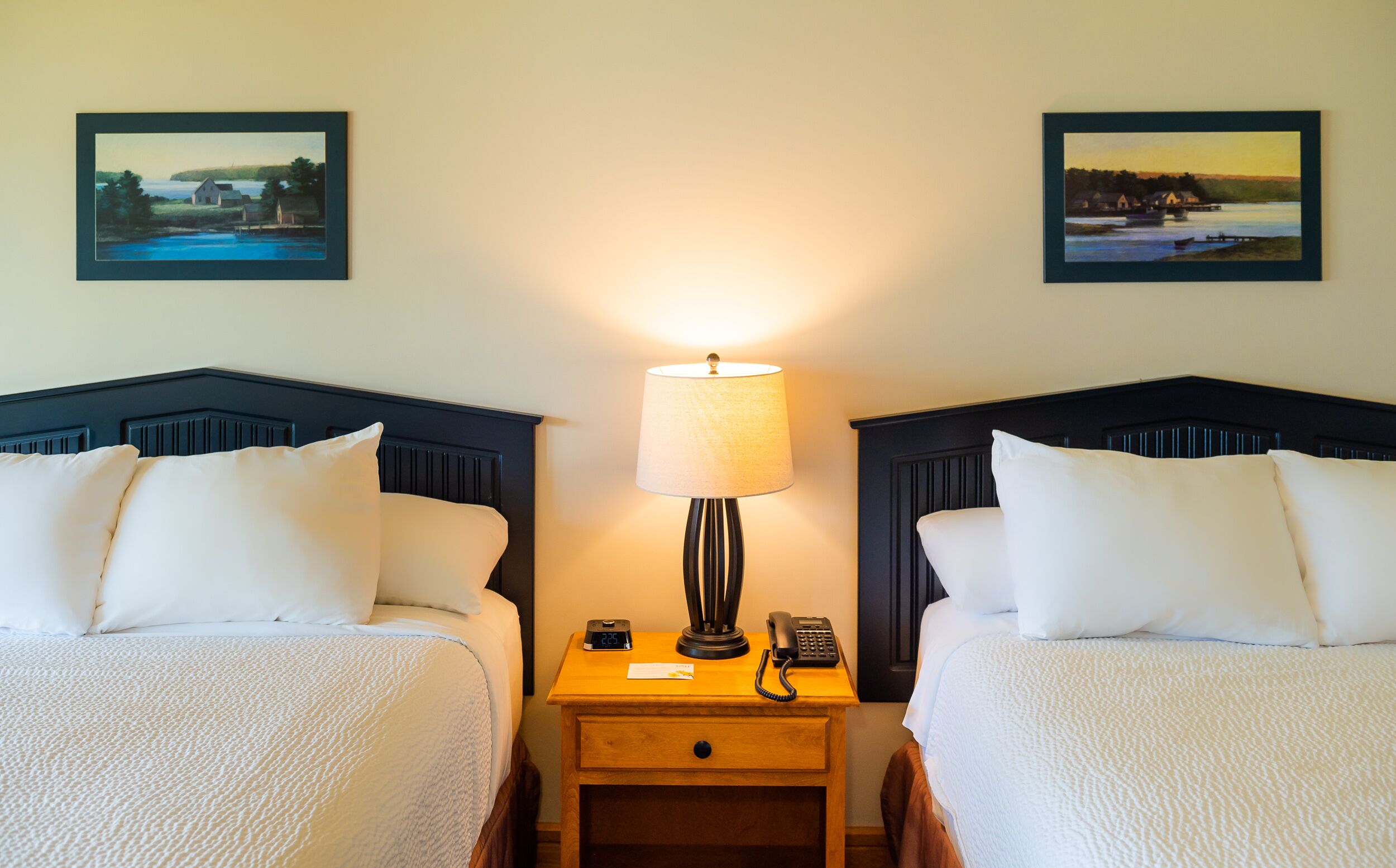 A hotel room with two beds, a nightstand with a lamp, a phone, and a small clock. There are two framed pictures on the wall.
