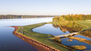 A golf course with a bridge over a calm lake, surrounded by trees and a clear sky.