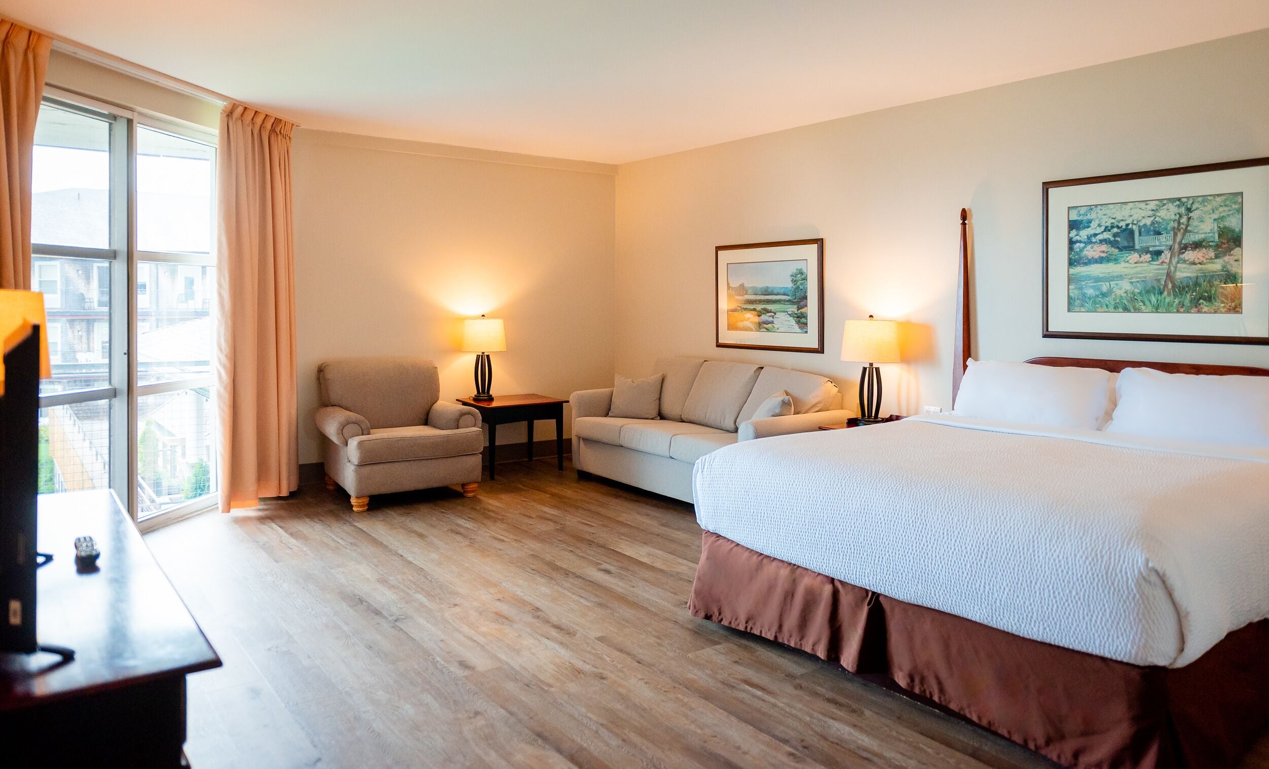 Habitación de hotel con cama, sofá, una silla y un escritorio. Hay dos lámparas sobre el escritorio y un cuadro en la pared.