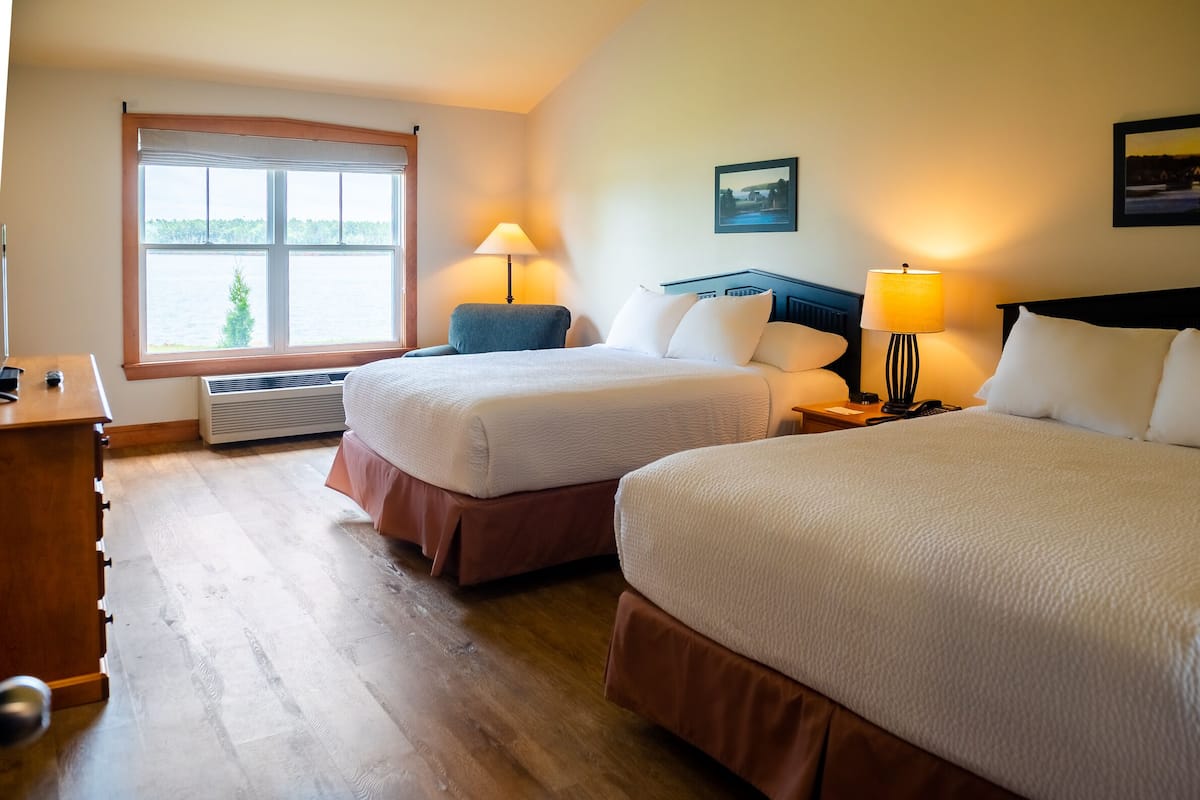 Habitación de hotel con dos camas, un escritorio, una silla, un ventanal con vistas a árboles y un cuadro en la pared.