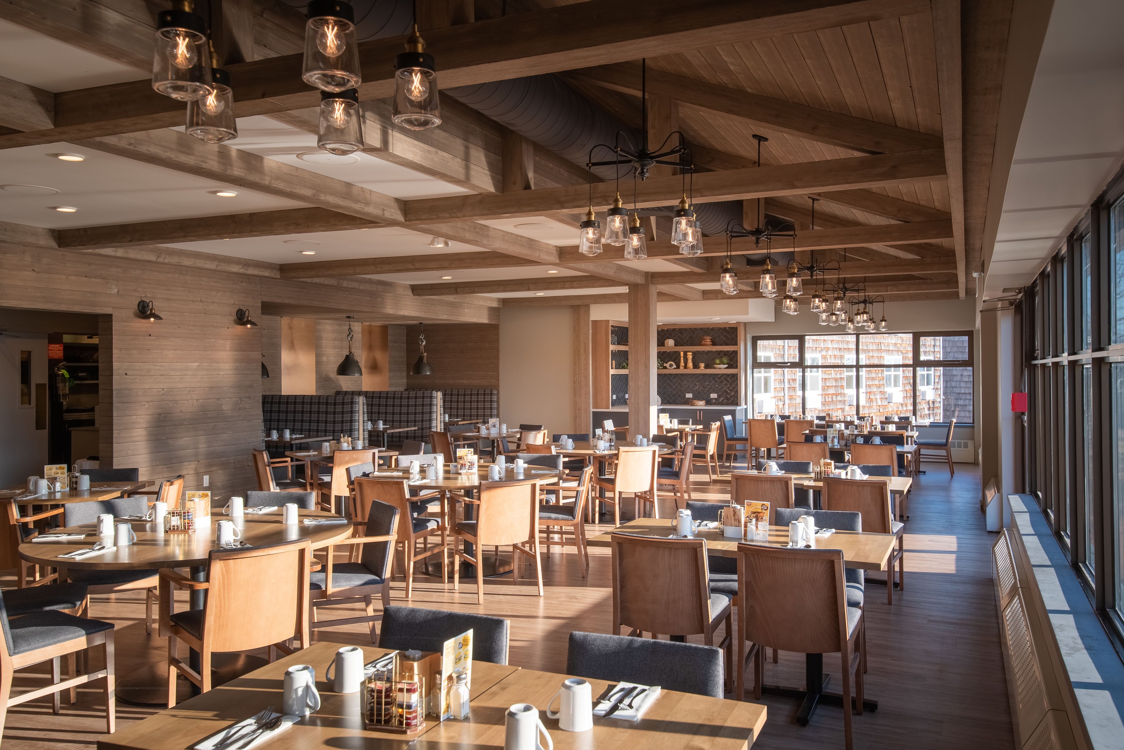 A spacious restaurant with wooden tables and chairs, hanging pendant lights, and large windows providing natural light.