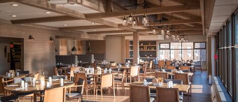 A spacious restaurant with wooden tables and chairs, hanging pendant lights, and large windows providing natural light.
