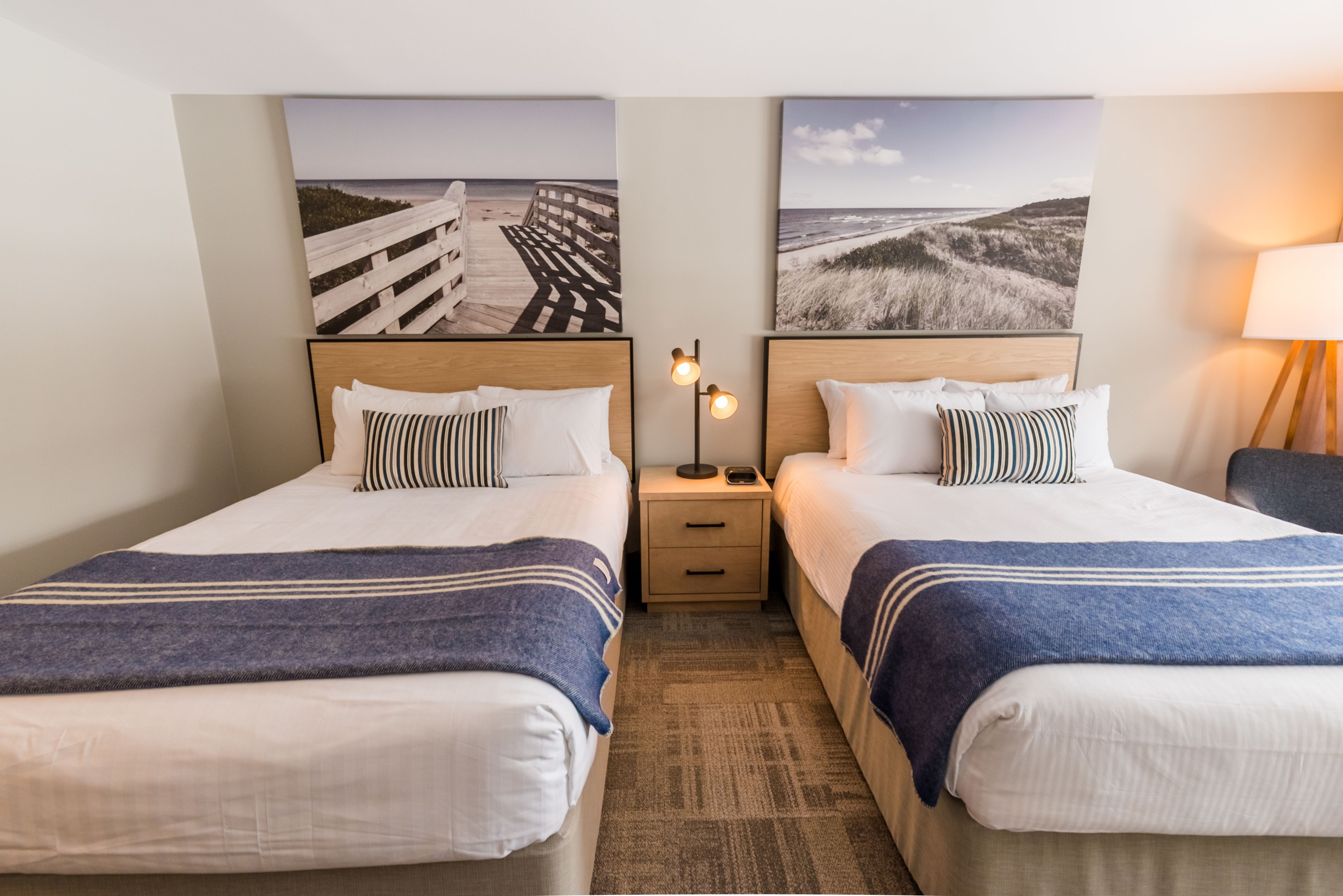 Una habitación de hotel con dos camas, cada una con cabecero de madera, una mesita de noche entre ellas y una lámpara a la derecha.