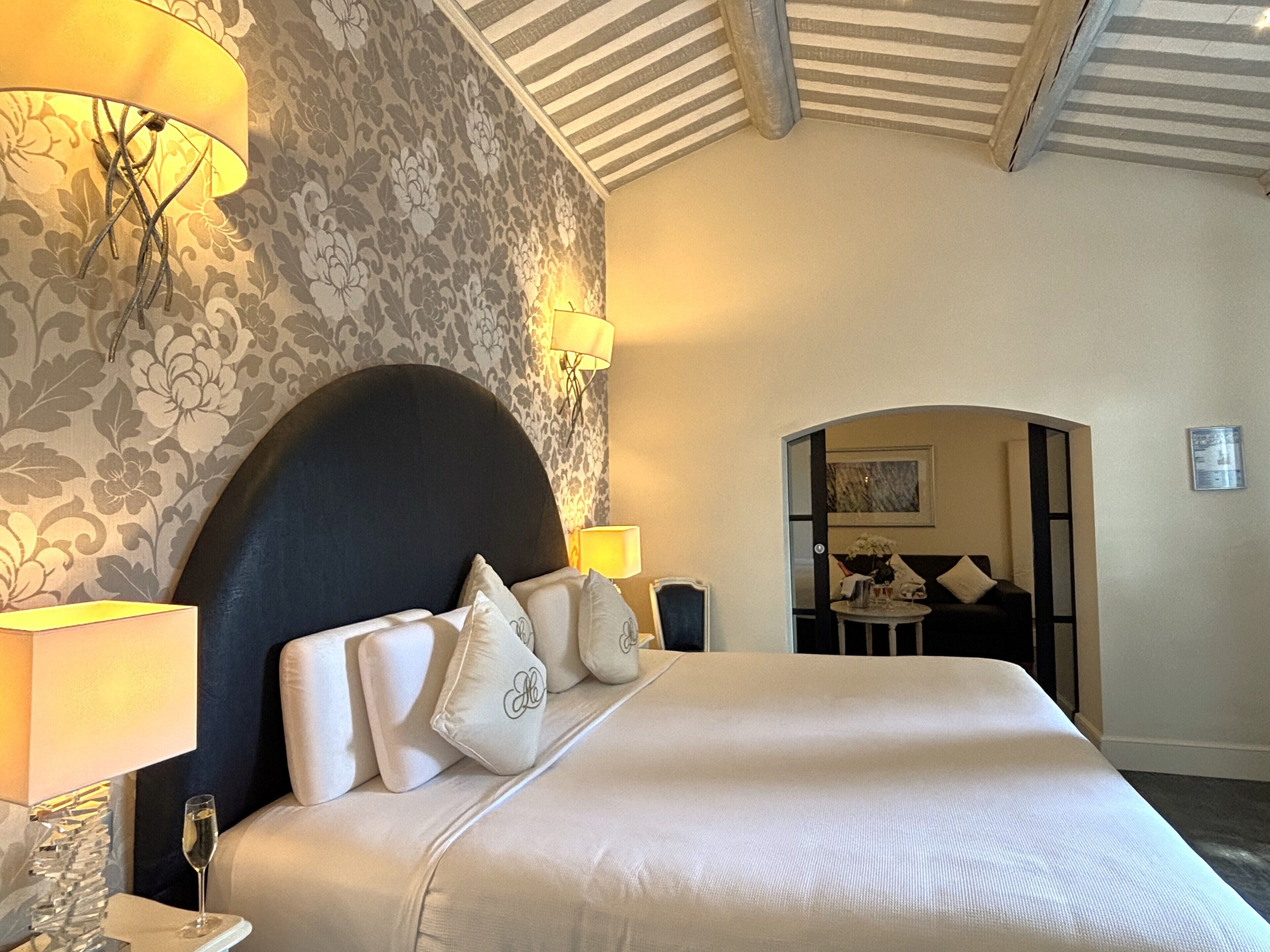 Een hotelkamer met een groot bed, twee bedlampjes, bloemenbehang en een uitzicht op een andere kamer door een boog.