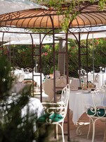 Un espacio de comedor al aire libre con mesas y sillas blancas, una columna central y una pérgola en la parte superior.