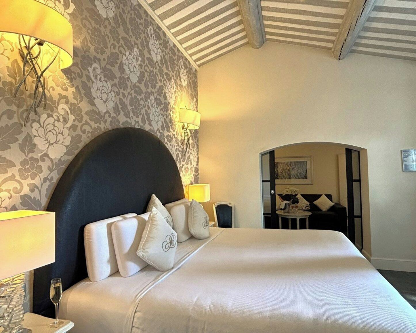A hotel room with a large bed, a headboard, and a floral wallpaper. There is a lamp on the left side of the bed and a small table with a lamp on the right.