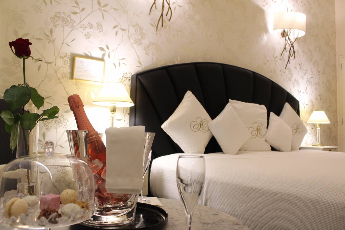 Une chambre d’hôtel avec un lit, une table de chevet, un verre d’eau, une bouteille de champagne et une rose sur la table.