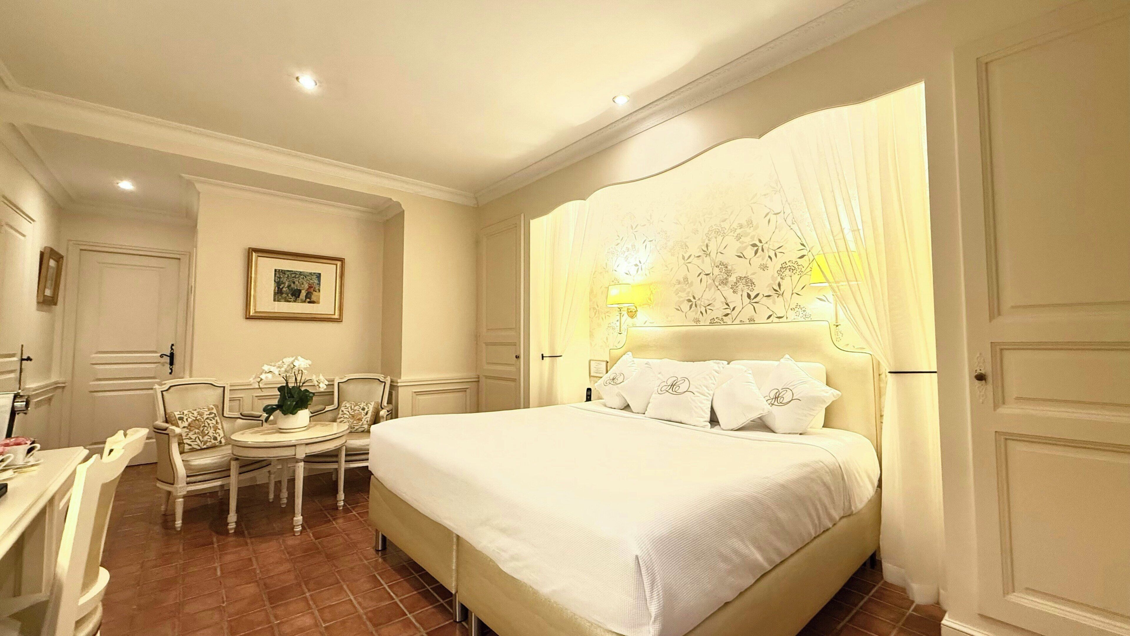 A hotel room with a bed, a chair, a small table with a plant, and a wall-mounted lamp.