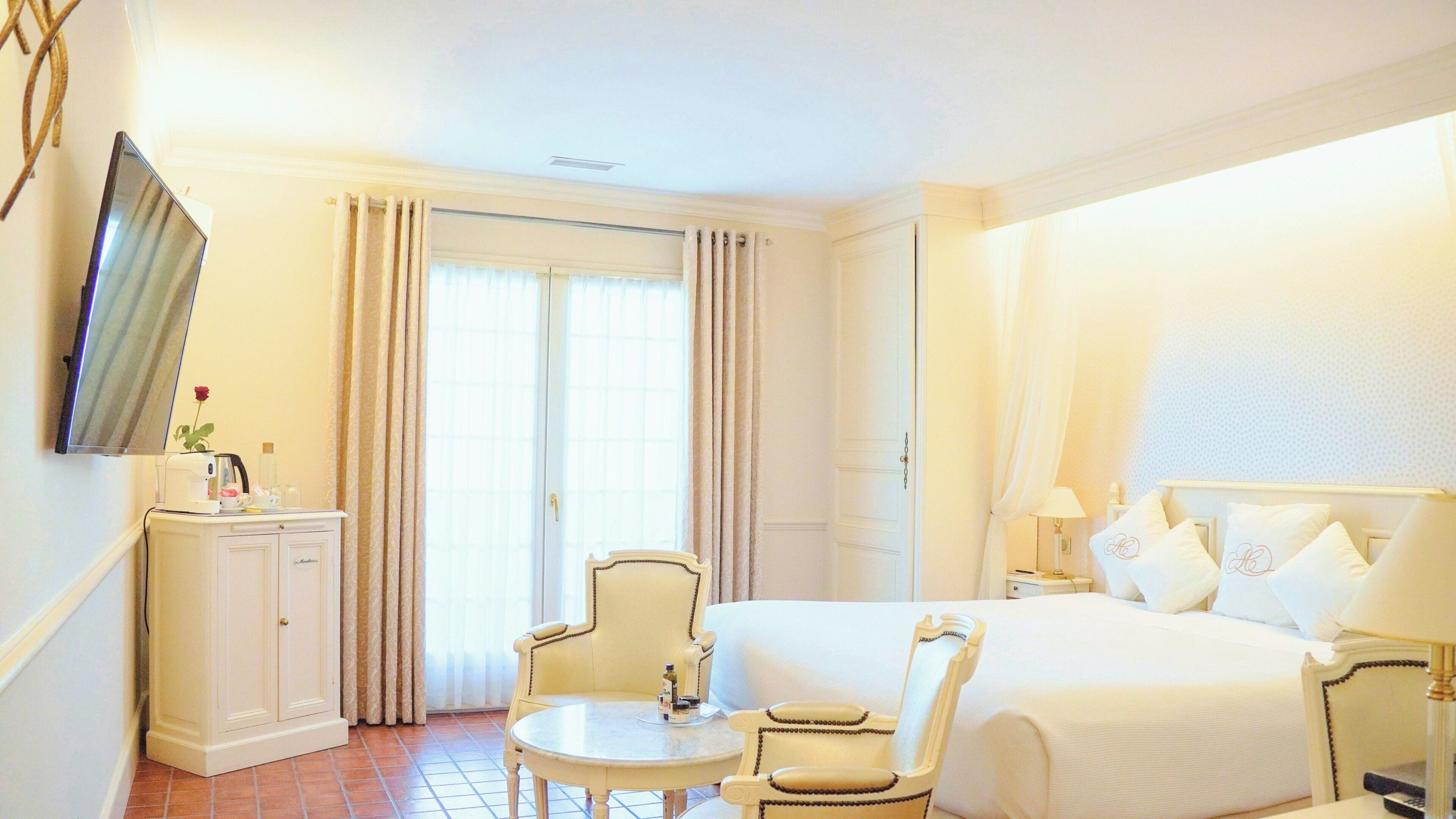 A hotel room with a bed, bedside tables, a chair, a small table, a TV, and a window with curtains.
