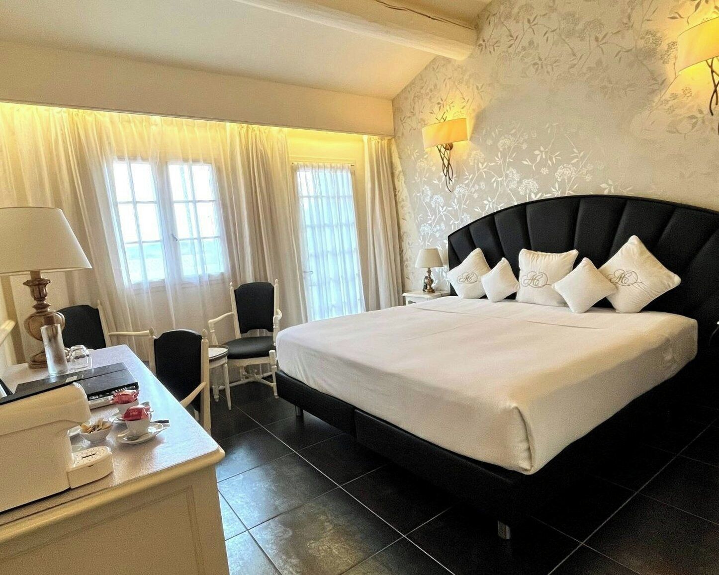 A hotel room with a large bed, a desk with a lamp, a chair, and a window with curtains.