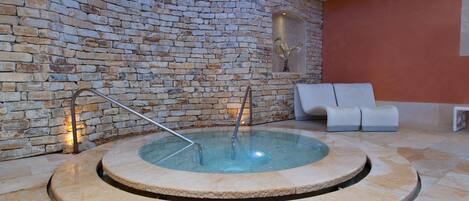 A stone-walled indoor pool area with a circular hot tub, a white bench, and a small table.