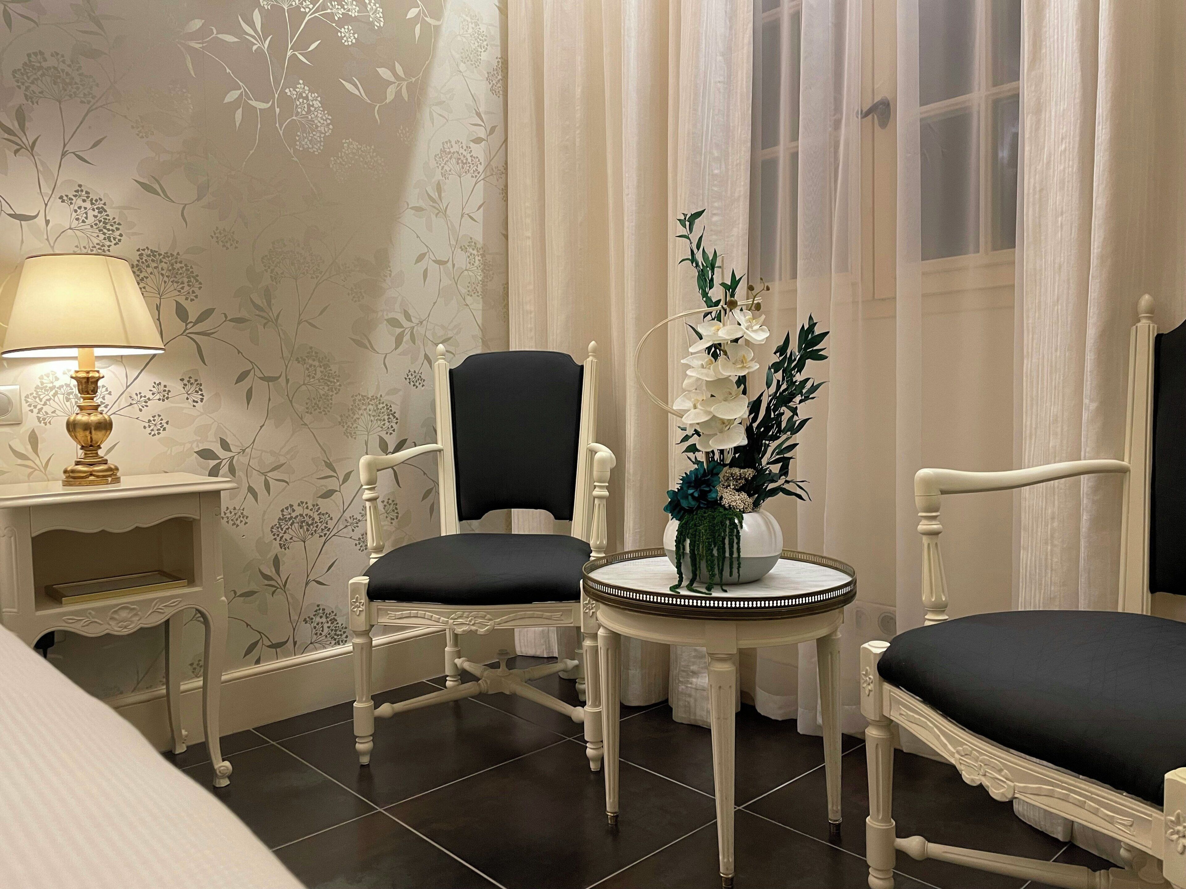 Une pièce comprenant un lit, deux chaises, une petite table avec un vase de fleurs, une lampe et une fenêtre avec des rideaux.