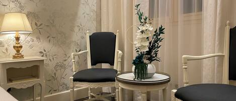 A room with a bed, two chairs, a small table with a vase of flowers, a lamp, and a window with curtains.