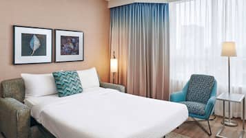 A hotel room with a bed, two framed leaf artworks, a chair, a lamp, and a view of the city through sheer curtains.