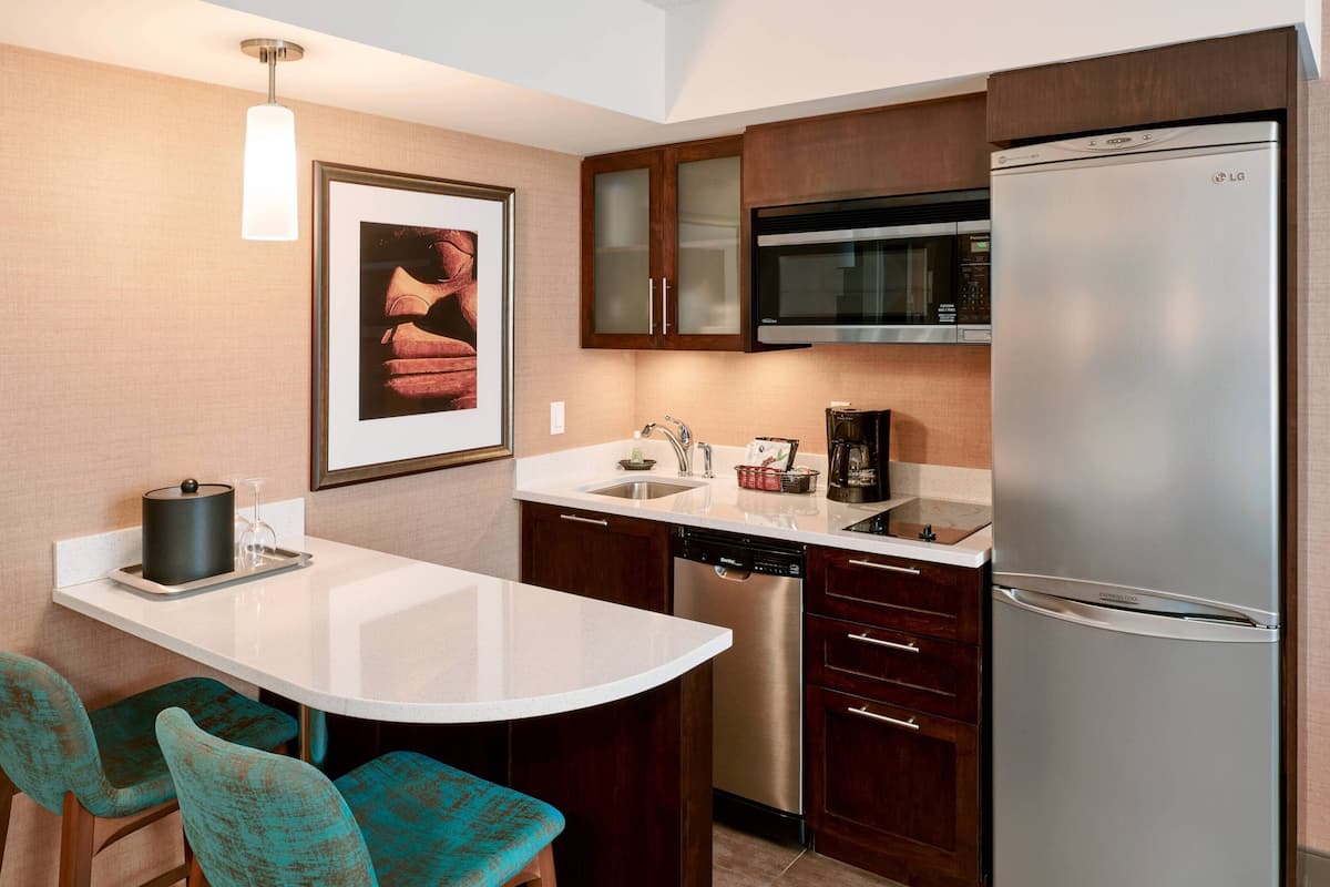 A modern kitchen with a stainless steel refrigerator, a microwave, and a dishwasher. There is a white countertop, wooden cabinets, and teal chairs.
