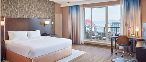 A hotel room with a large bed, a desk with a chair, a small table, and a balcony with a view of buildings.