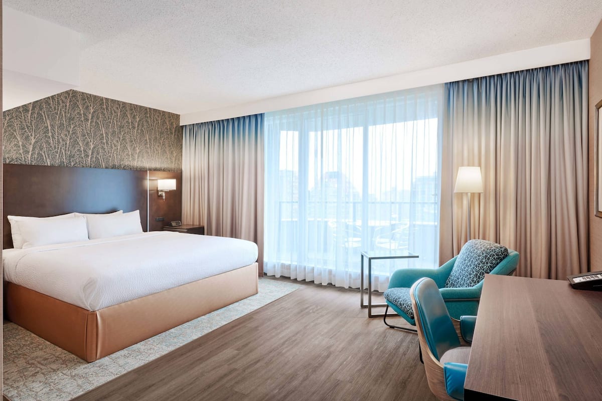A hotel room with a large bed, a desk, a chair, a lamp, and a view of the city through large windows.