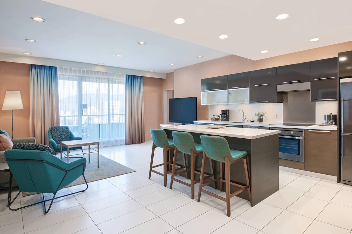 A modern kitchen with a central island, bar stools, a dining area with a table and chairs, and a large window with curtains.
