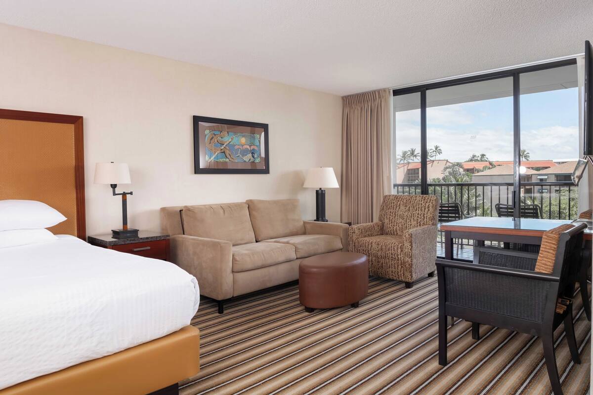 Habitación de hotel con cama, sofá, sillón, una mesita y un balcón con vistas a palmeras y edificios.