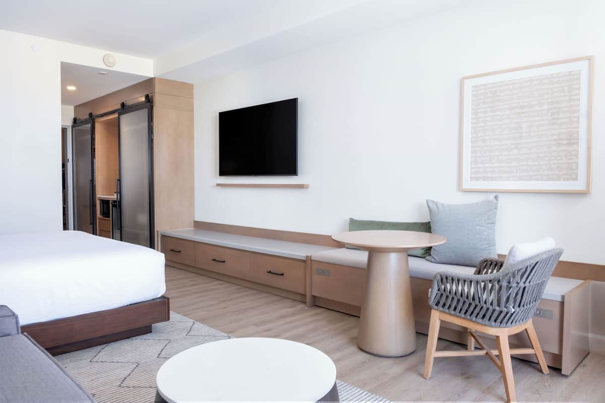 Habitación de hotel moderna con cama, sofá, una mesita, televisor y un cuadro colgado en la pared.