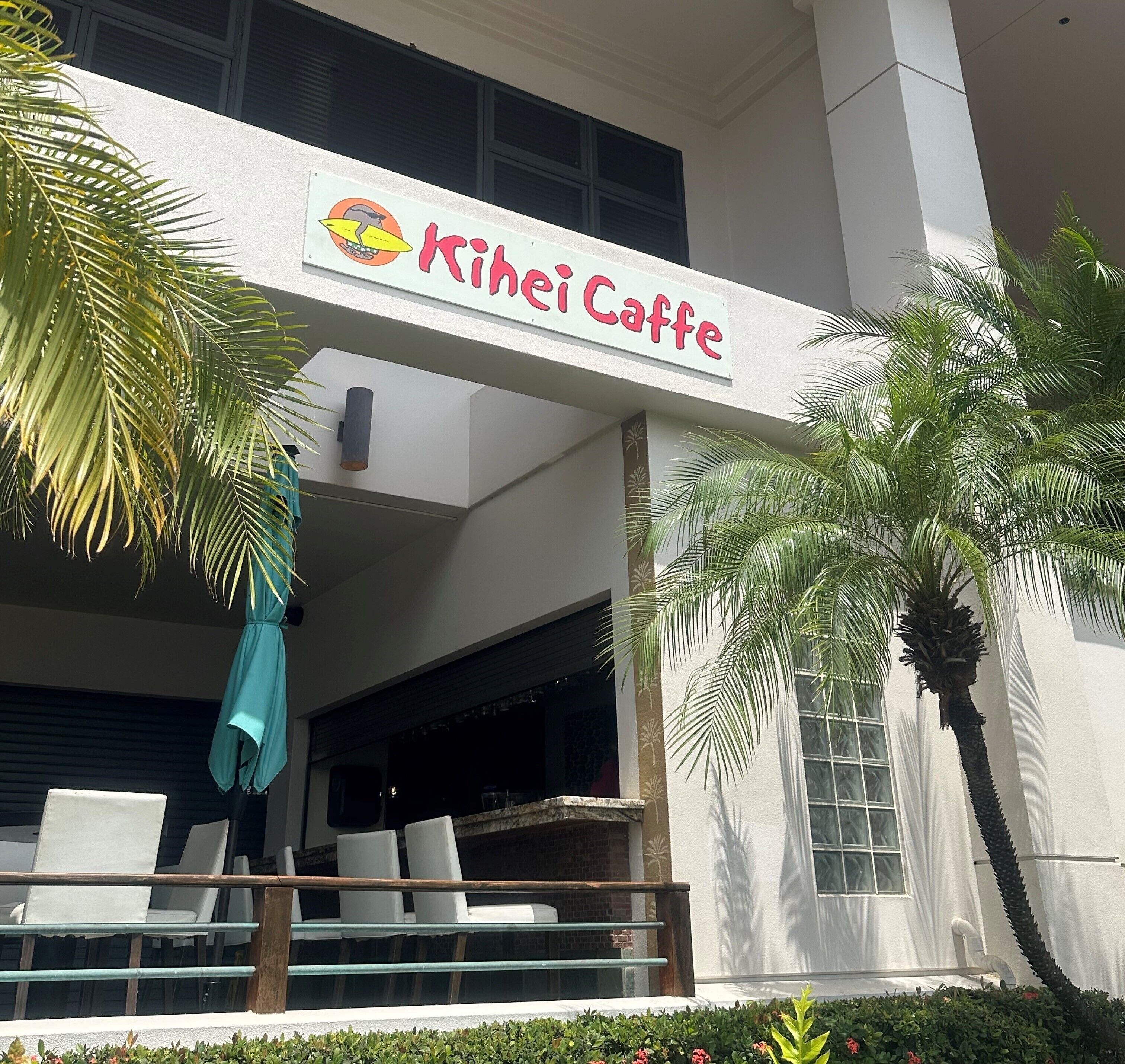 A building with a sign reading "Kihei Caffe" and a covered outdoor seating area with chairs and a table.