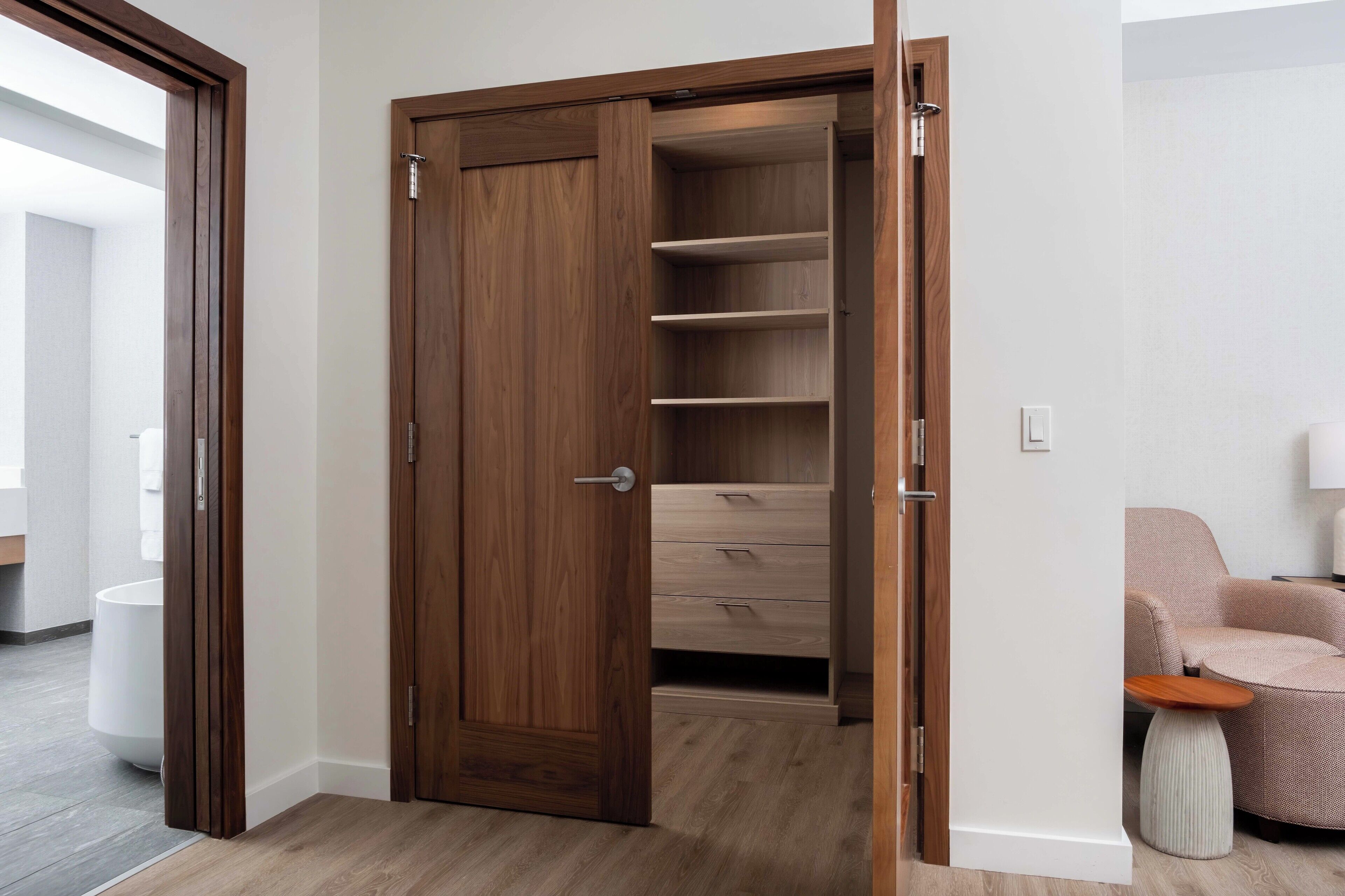 A wooden wardrobe with multiple shelves and drawers, a small round table, and a chair in a modern room with a light-colored floor and white walls.