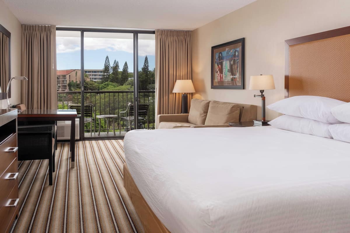 Habitación de hotel con una cama grande, un escritorio, una silla, un balcón con vistas a zonas verdes y edificios, y un cuadro en la pared.