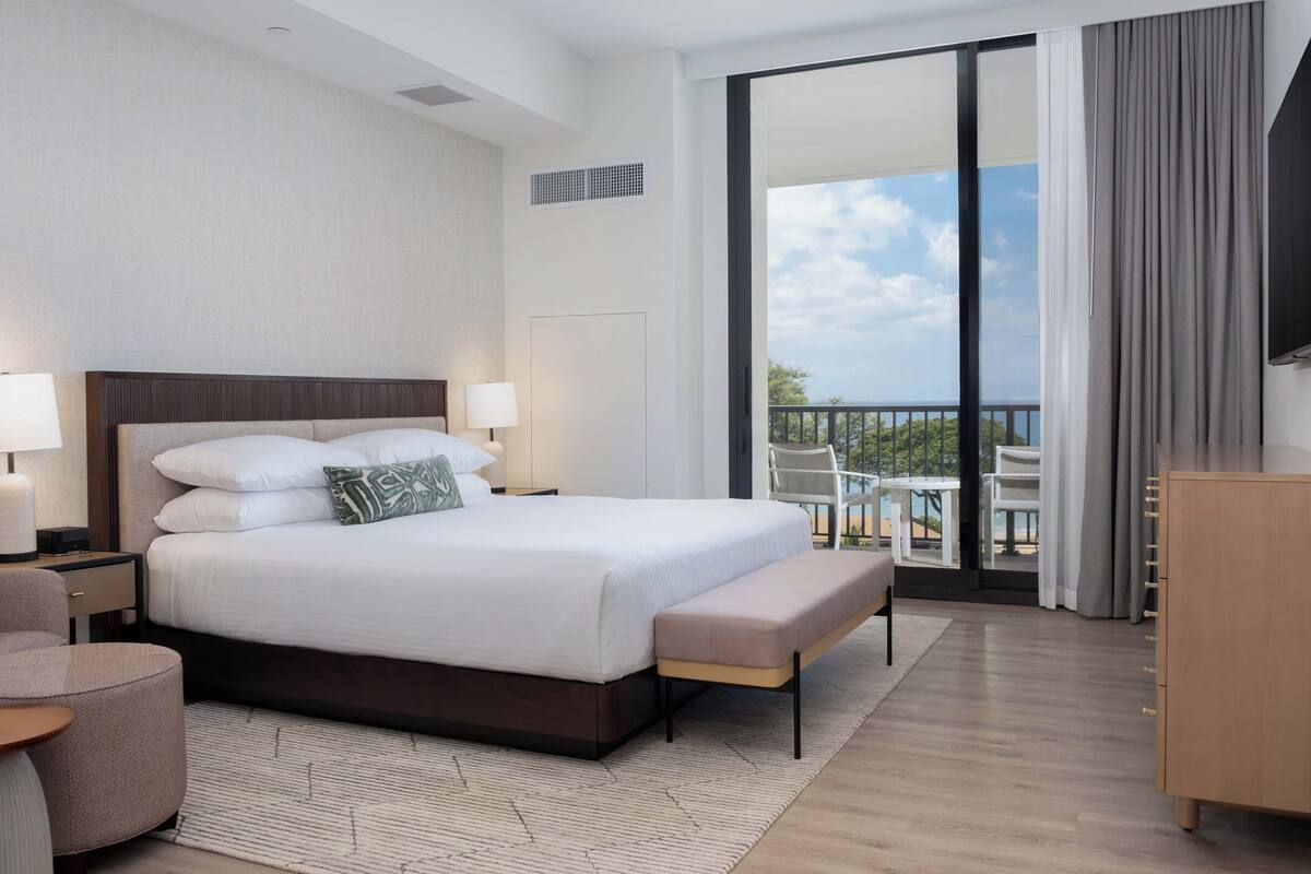 Una habitación de hotel moderna con una cama grande, lámparas de noche, un banco, una mesita y vistas al exterior a través de una puerta corredera de vidrio.