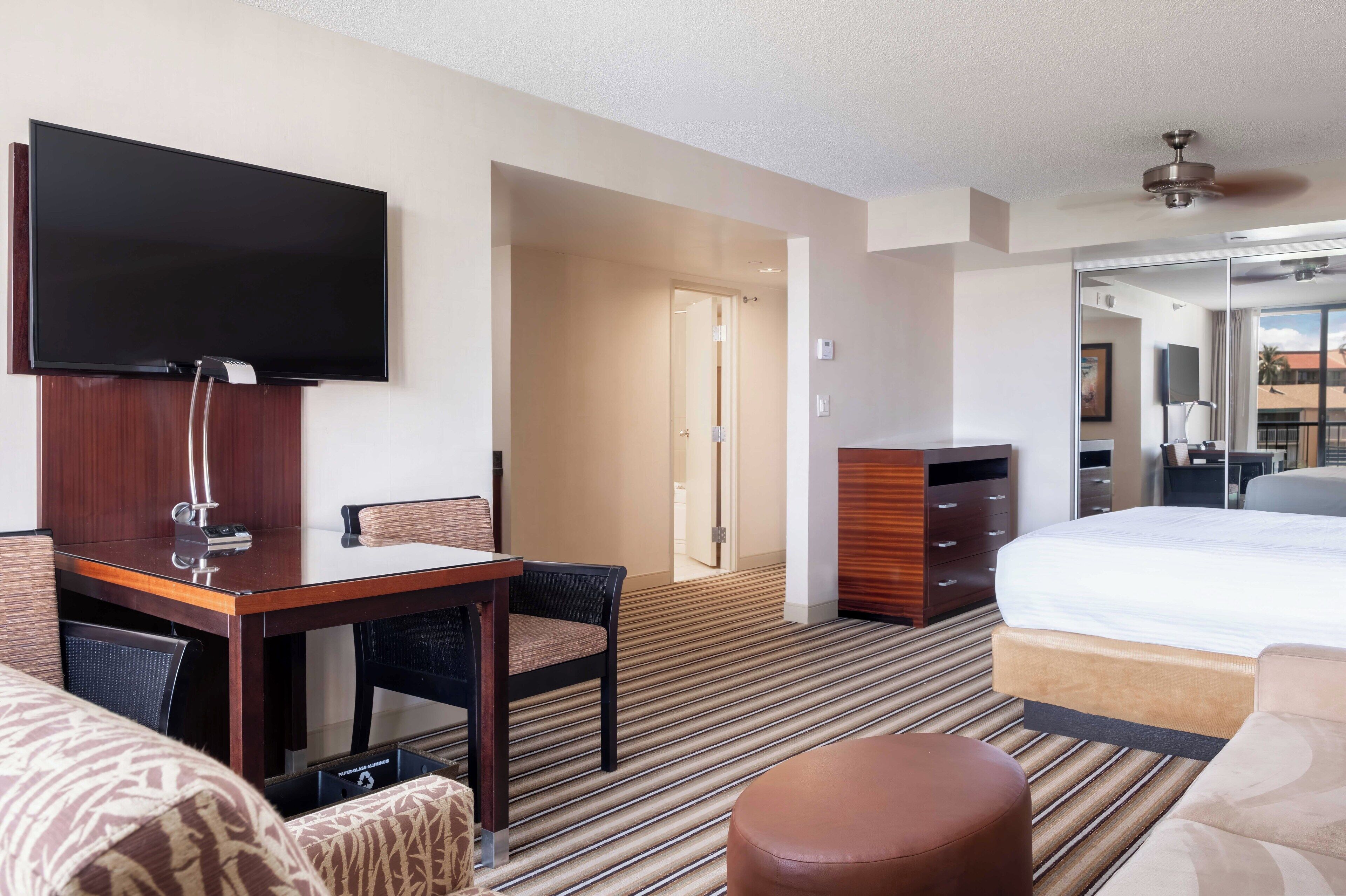 A hotel room with a flat-screen TV, a wooden desk, a bed, a sofa, and a bathroom visible through an open door.