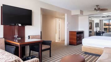 A hotel room with a flat-screen TV, a wooden desk, a bed, a sofa, and a bathroom visible through an open door.