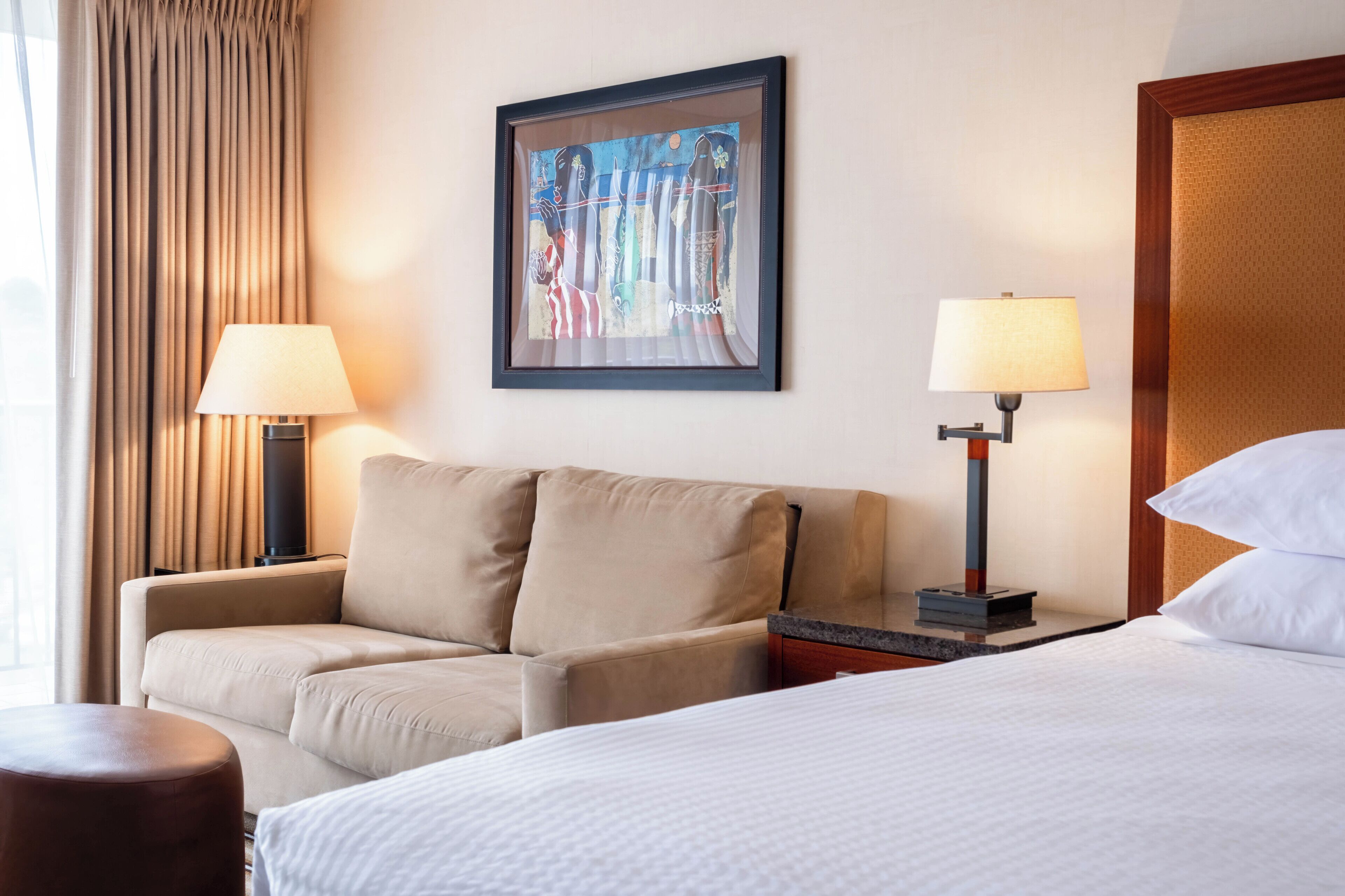 A hotel room with a bed, a sofa, two lamps, a painting on the wall, and a window with curtains.