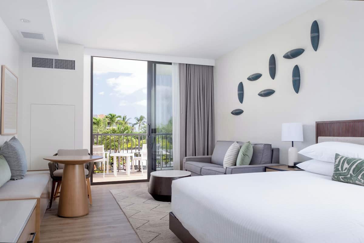 Habitación de hotel con cama, sofá, mesa de comedor y balcón con vista a palmeras.